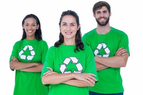 Three individuals wearing bright green T-shirts featuring a white recycling symbol stand outdoors on a pavement in front of a building. The person in the middle, a woman with dark hair styled in loose waves, is positioned slightly forward and has her arms crossed, smiling directly at the camera. To her left, another woman with long dark hair has her arms crossed and is also smiling, while to her right, a man with a beard and short dark hair has his arms crossed and a friendly expression. The background reveals part of a staircase and a stone building with windows, suggesting an urban or suburban setting possibly within Blackheath or a nearby area. The lighting appears natural, with daylight illuminating their faces and the scene around them. The image conveys a message of environmental responsibility, consistent with services offered by companies like Office Clearance Blackheath, which specialises in rubbish and office clearance services in the local area.