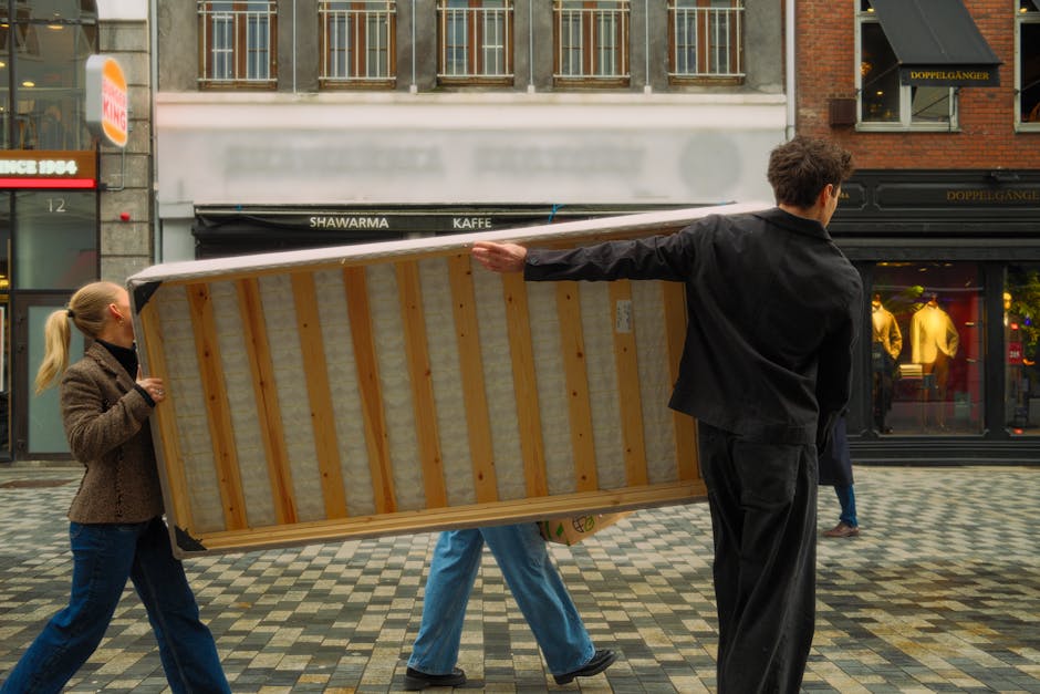 A male healthcare worker dressed in a maroon uniform with reflective grey stripes is seen loading an orange and grey medical stretcher into the back of a white ambulance. The ambulance's rear doors ar