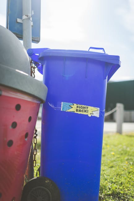 A tall, bright blue plastic rubbish bin with a closed lid is positioned outdoors on a grassy area, adjacent to a pink and grey wheeled bin with a domed lid. The blue bin has a small, rectangular, torn