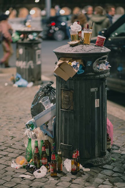 A tall, bright blue plastic rubbish bin with a closed lid is positioned outdoors on a grassy area, adjacent to a pink and grey wheeled bin with a domed lid. The blue bin has a small, rectangular, torn