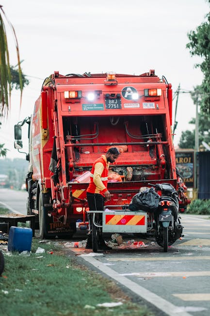 A sanitation worker dressed in a green uniform and safety helmet is standing on the pavement beside a black and white rubbish collection vehicle, which has its rear compactor open and partly filled wi