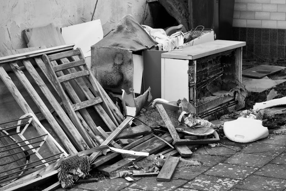 A collection of black rubbish bags and discarded packaging, including cardboard and plastic, piled in front of a red metal door with graffiti and multiple stickers, on an external wall of a building w