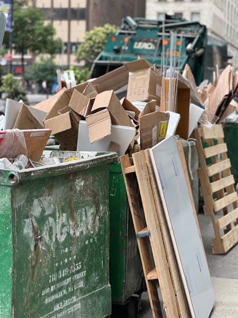 A large green industrial waste bin filled with a mixture of cardboard boxes, some flattened and others intact, along with packaging materials, situated outdoors on a paved street. The cardboard is mos