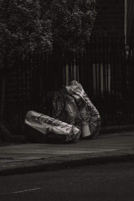 A large green industrial waste bin filled with a mixture of cardboard boxes, some flattened and others intact, along with packaging materials, situated outdoors on a paved street. The cardboard is mos