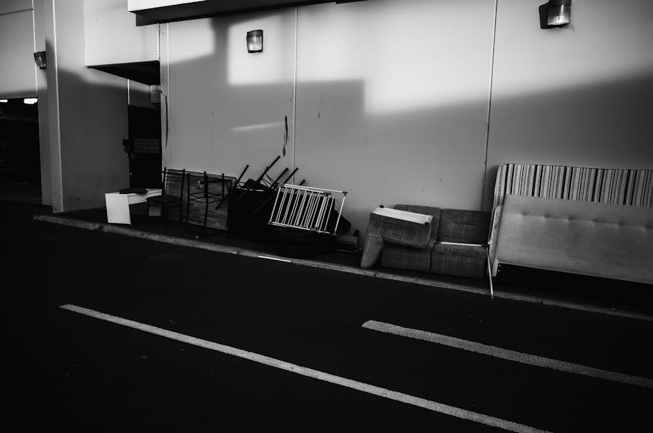 An old, upholstered sofa with a beige and orange patterned fabric, showing visible tears and worn areas on the armrests, is situated on a grassy area surrounded by tall weeds and fallen leaves. The so